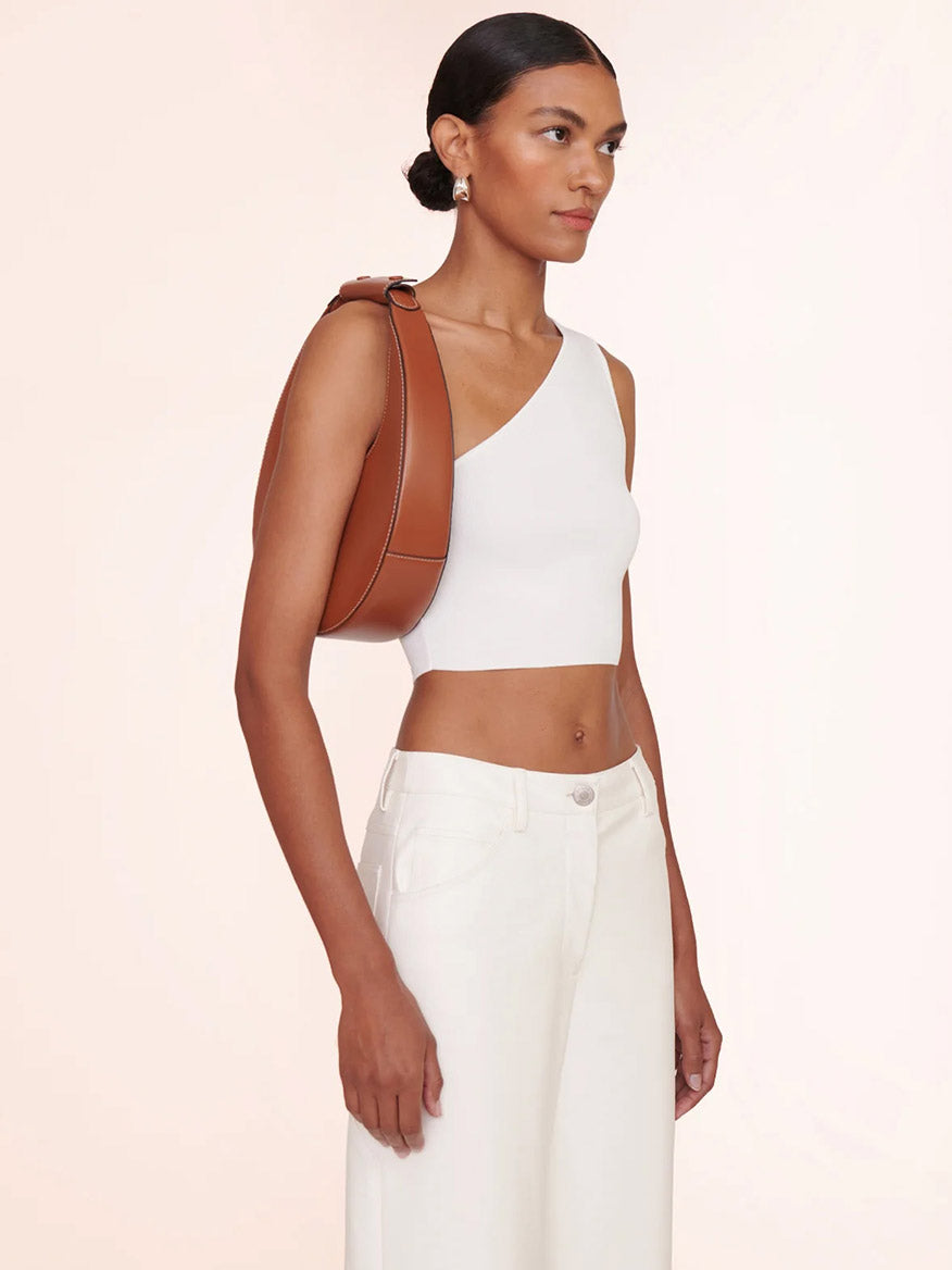 Carrying the STAUD Moon Tote Bag in Tan, a person wears a white crop top and high-waisted pants. Styled with earrings and hair tied back, they stand gracefully against a plain background.