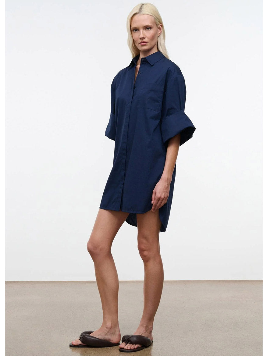 A person stands indoors wearing the STAUD Nico Dress in Navy, an oversized cotton poplin button-down shirt dress, paired with dark slide sandals and arms relaxed at their sides.
