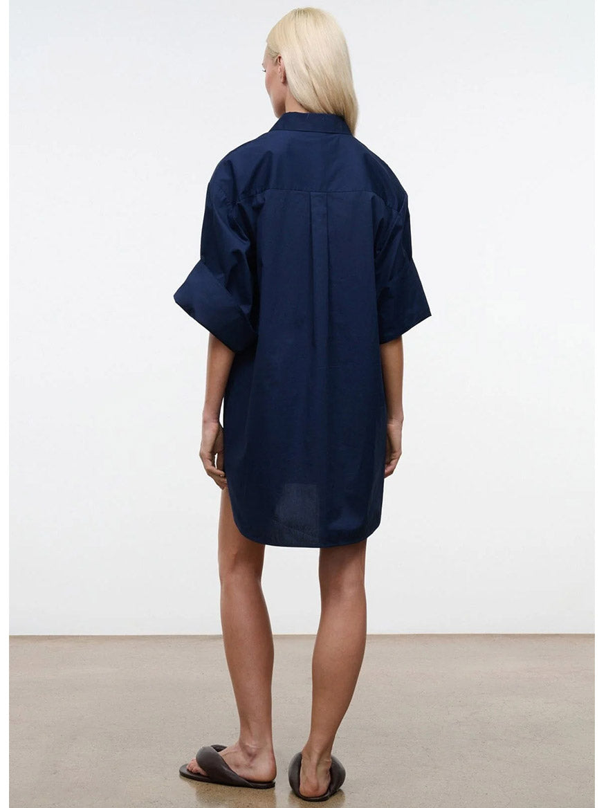 A person with long blonde hair stands facing away, wearing the STAUD Nico Dress in Navy—a navy oversized mini shirtdress with adjustable sleeves—and black slide sandals, set against a plain light background.
