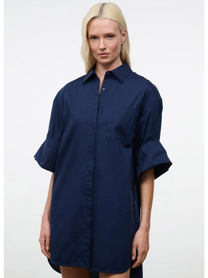 A person with long blonde hair wears the STAUD Nico Dress in Navy, an oversized cotton poplin button-up dress with adjustable sleeves, standing against a plain white background.