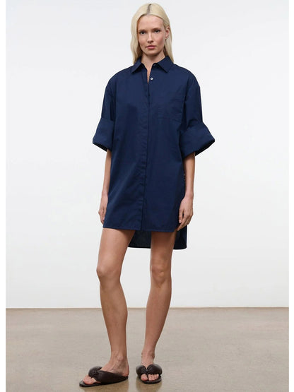A person stands indoors against a plain background, wearing the STAUD Nico Dress in Navy—an oversized mini shirtdress with adjustable sleeves—paired with dark sandals.