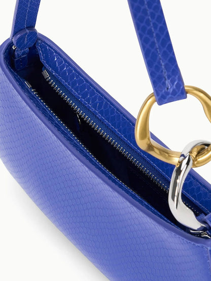 A close-up of the STAUD Ollie Bag in Cobalt shows its blue textured snake-print leather, zipper closure, and mixed-metal hardware on the strap.