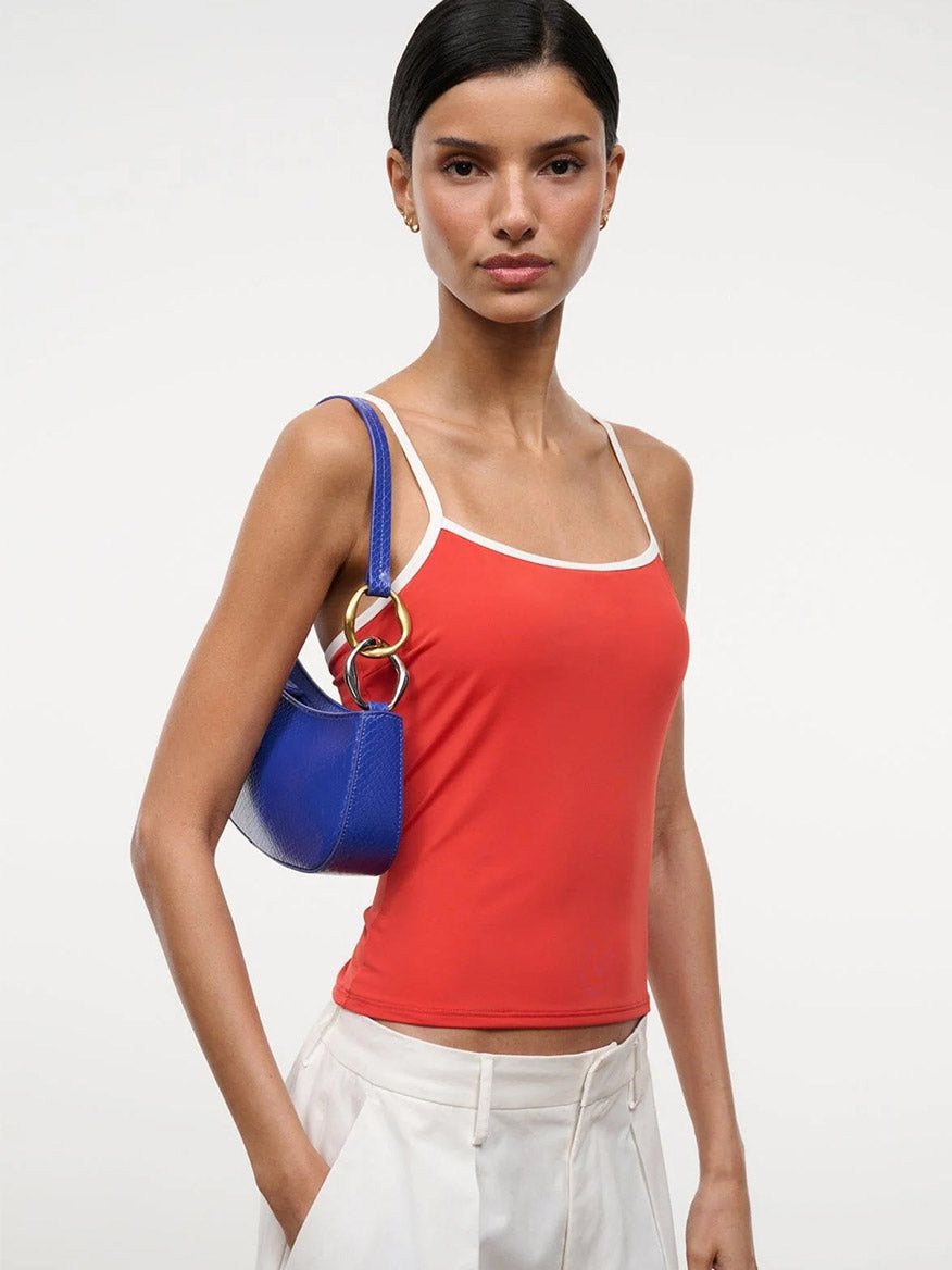 A woman in a red camisole and white pants holds the STAUD Ollie Bag in Cobalt, featuring mixed-metal hardware, over her shoulder against a plain white background.