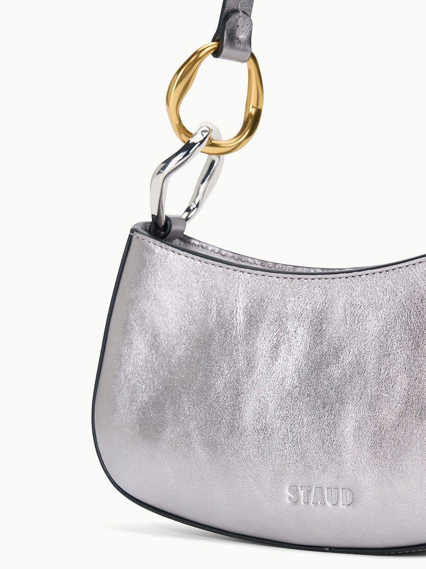 The STAUD Ollie Bag in Pewter is a metallic leather mini shoulder bag with a curved shape and two-tone gold and silver hardware on the handle, shown against a plain background.