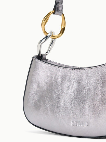 The STAUD Ollie Bag in Pewter is a metallic leather mini shoulder bag with a curved shape and two-tone gold and silver hardware on the handle, shown against a plain background.