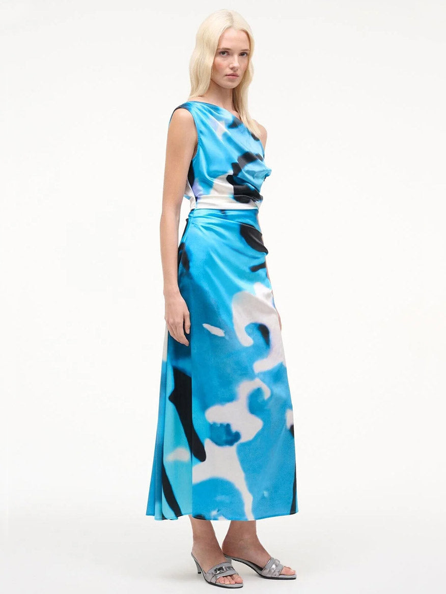A woman poses against a plain white background wearing the STAUD Phare Silk Dress in Blue Haze, a sleeveless, ankle-length abstract print special occasion dress, paired with silver heeled sandals.