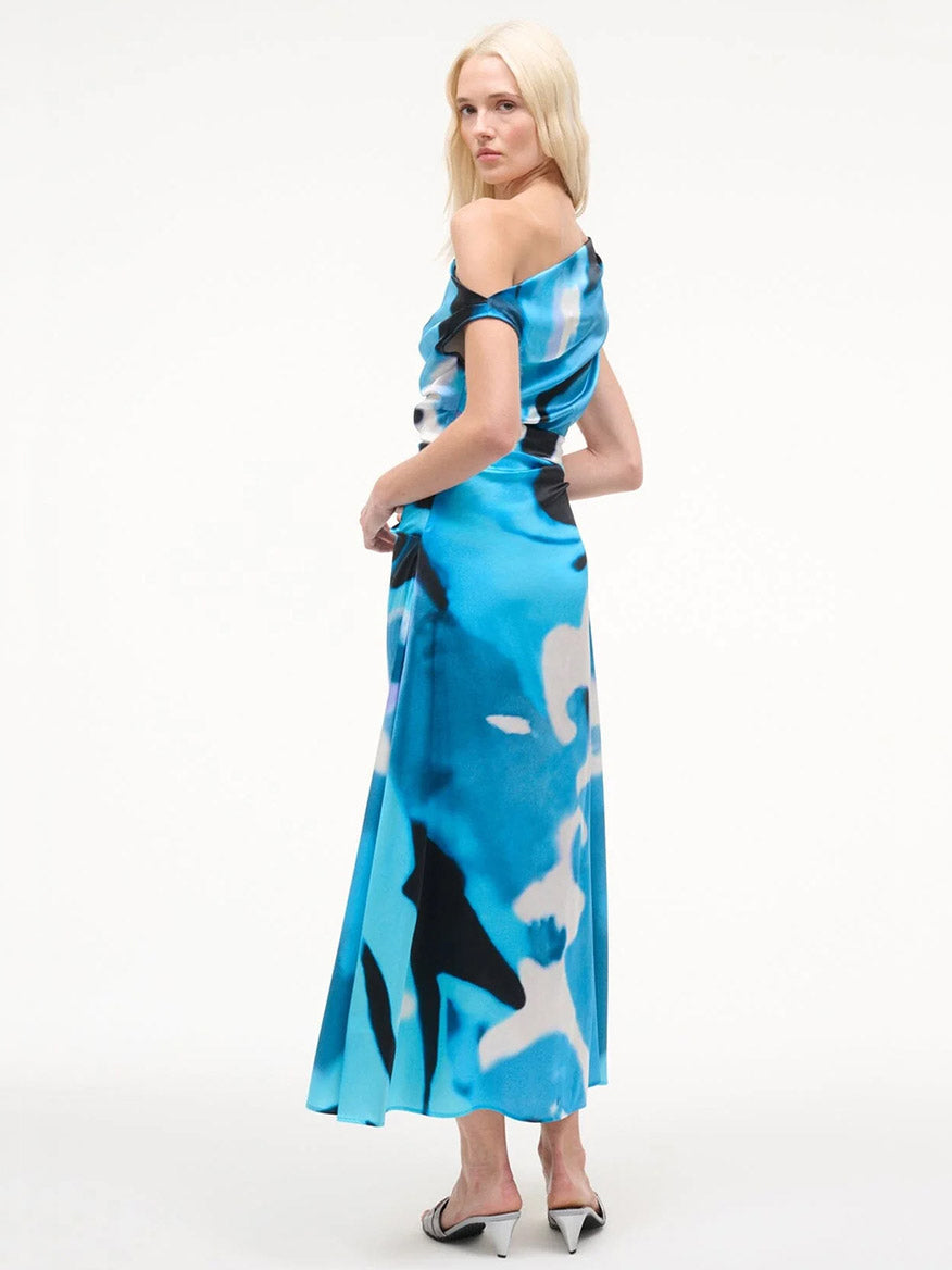 A woman stands in a three-quarter pose against a plain white background, wearing the STAUD Phare Silk Dress in Blue Haze with white heels—perfect for special occasions.
