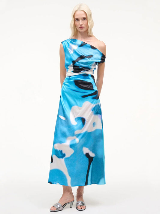 A woman poses against a white background wearing the STAUD Phare Silk Dress in Blue Haze, an off-the-shoulder, abstract-printed special occasion dress, styled with silver open-toe sandals.