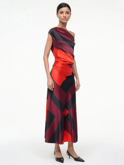 A woman stands in the STAUD Phare Silk Dress in Ruby Prism, an ankle-length, one-shoulder stretch silk dress featuring bold red and black geometric patterns, paired with black heels against a plain white background.