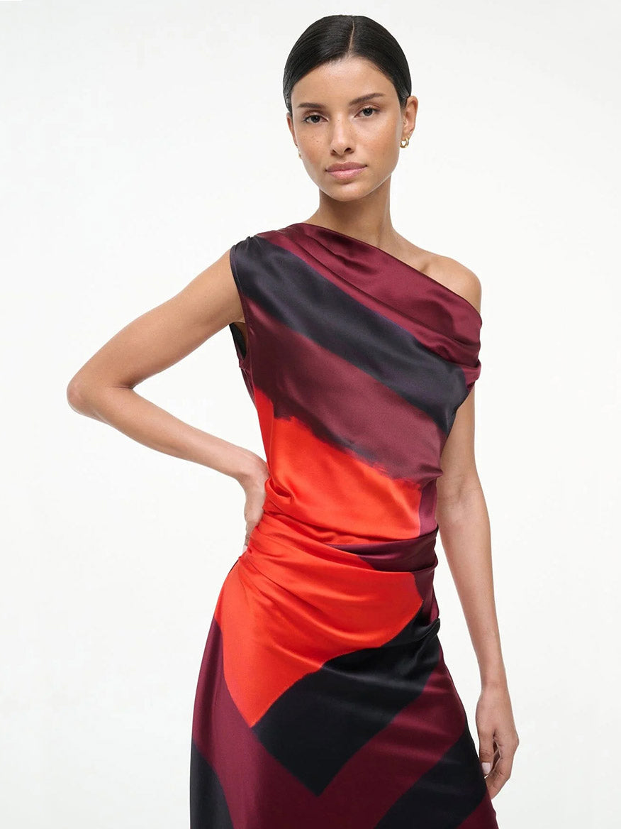 A woman stands against a plain background wearing the STAUD Phare Silk Dress in Ruby Prism—a one-shoulder special occasion dress with a striking red and black geometric pattern.