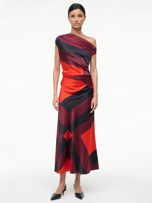 A woman stands against a plain background wearing the STAUD Phare Silk Dress in Ruby Prism, an off-the-shoulder, ankle-length special occasion dress with a bold red and black abstract pattern and black pointed shoes.