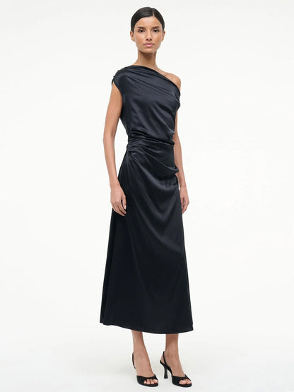 A woman stands against a plain background wearing the STAUD Phare Silk Dress in Black, a one-shoulder, ankle-length stretch silk dress with ruched detailing, paired with black open-toe heels.