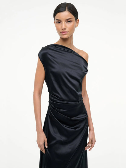 A woman stands against a plain background wearing the STAUD Phare Silk Dress in Black—a sleek, off-the-shoulder design with draped waist detailing perfect for special occasions.