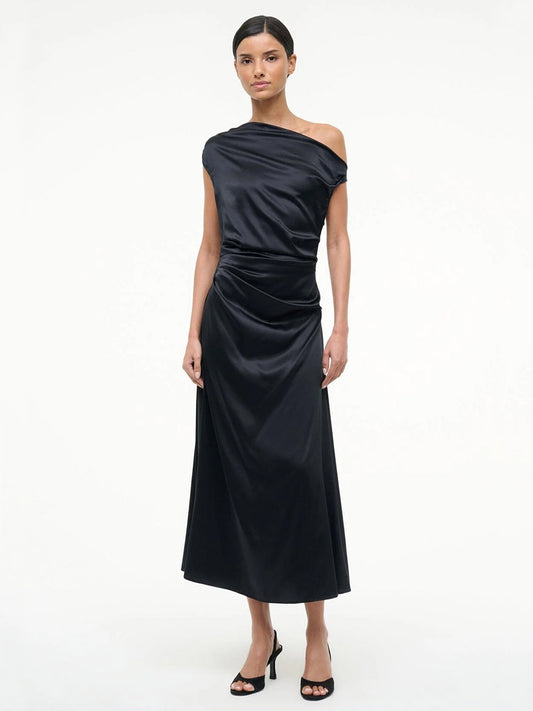 A woman stands against a plain background wearing the STAUD Phare Silk Dress in Black, featuring a sleek one-shoulder design, paired with black high-heeled sandals—a stunning choice for any special occasion.