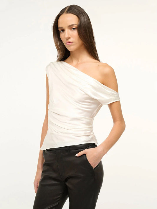A woman stands against a plain background wearing the STAUD Phare Silk Top in Ivory, an off-the-shoulder, drapey silhouette paired with black pants, her hand casually in her pocket.