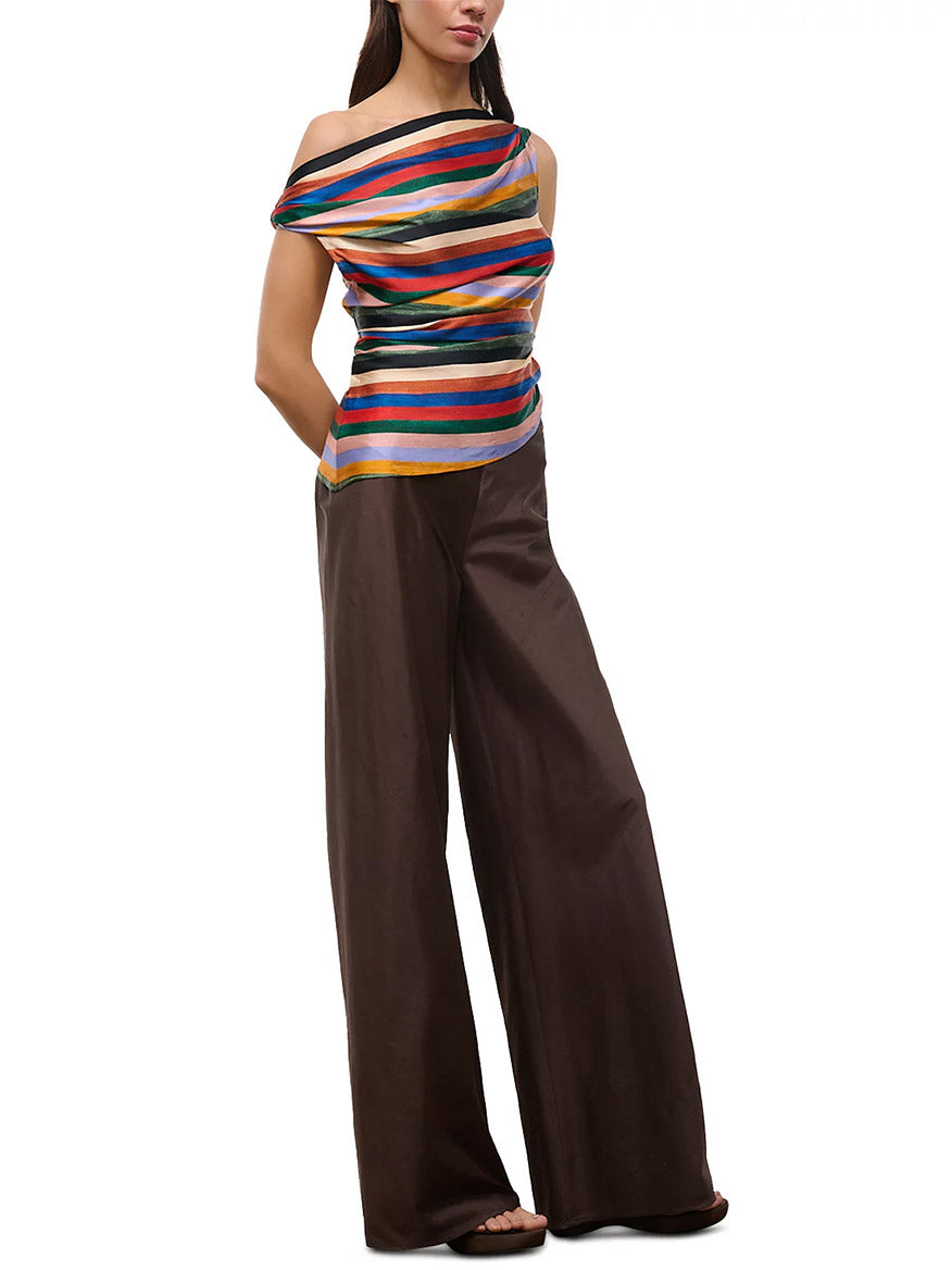 Woman wearing a colorful STAUD Phare Silk Top in Artist Stripe and wide-leg brown pants, standing with hands behind her back against a white background.