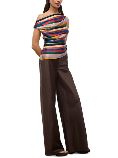 Woman wearing a colorful STAUD Phare Silk Top in Artist Stripe and wide-leg brown pants, standing with hands behind her back against a white background.