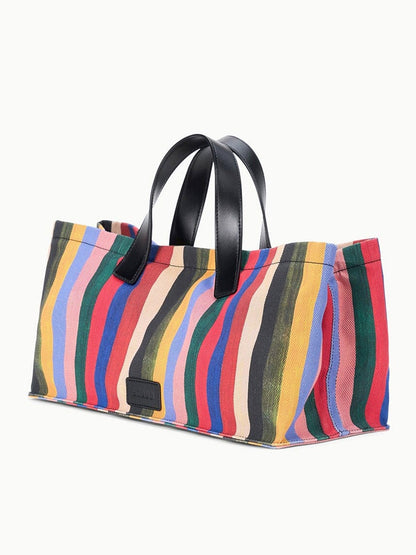The STAUD Port Tote in Artist Stripe is a rectangular printed canvas bag with black leather handles, showcasing a vibrant wavy striped pattern in red, blue, green, yellow, and purple.