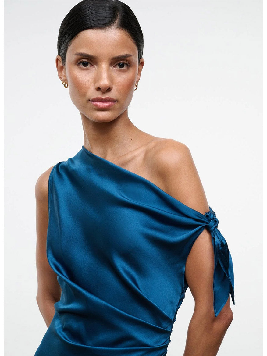 A woman with dark hair styled in a low bun wears the STAUD Portia Silk Dress in Harbor—an asymmetrical one-shoulder silk dress featuring an adjustable tie detail on the right shoulder—while standing against a plain light background.