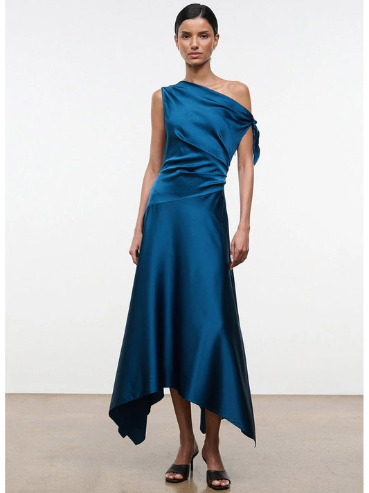 A woman stands against a plain background wearing the STAUD Portia Silk Dress in Harbor—a teal, one-shoulder asymmetrical silk dress with an adjustable tie detail and a midi-length skirt—paired with black open-toe heels.