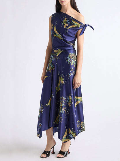 A woman wearing the STAUD Portia Silk Dress in Tapestry Midnight—a navy blue, one-shoulder dress featuring an asymmetrical hemline and adorned with intricate tapestry-inspired patterns of birds and blooms—stands against a plain white background.