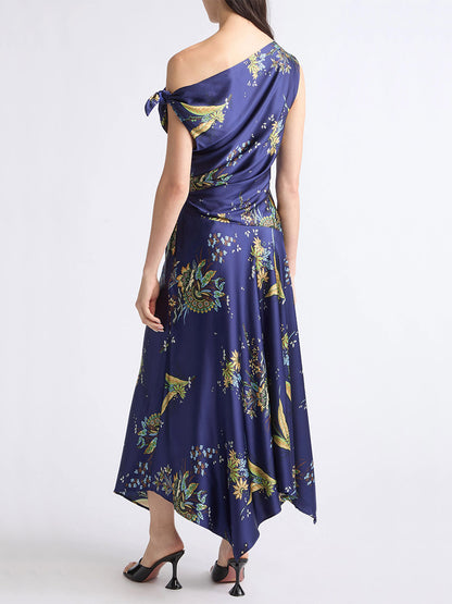 A woman stands with her back to the camera, wearing a flowery printed one-shoulder dress with an asymmetrical hem and black-heeled sandals.