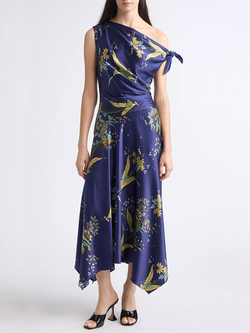 A woman wearing the STAUD Portia Silk Dress in Tapestry Midnight—a navy blue, one-shoulder dress featuring an asymmetrical silhouette and a delicate tapestry-inspired print of florals and birds—stands against a plain white background.