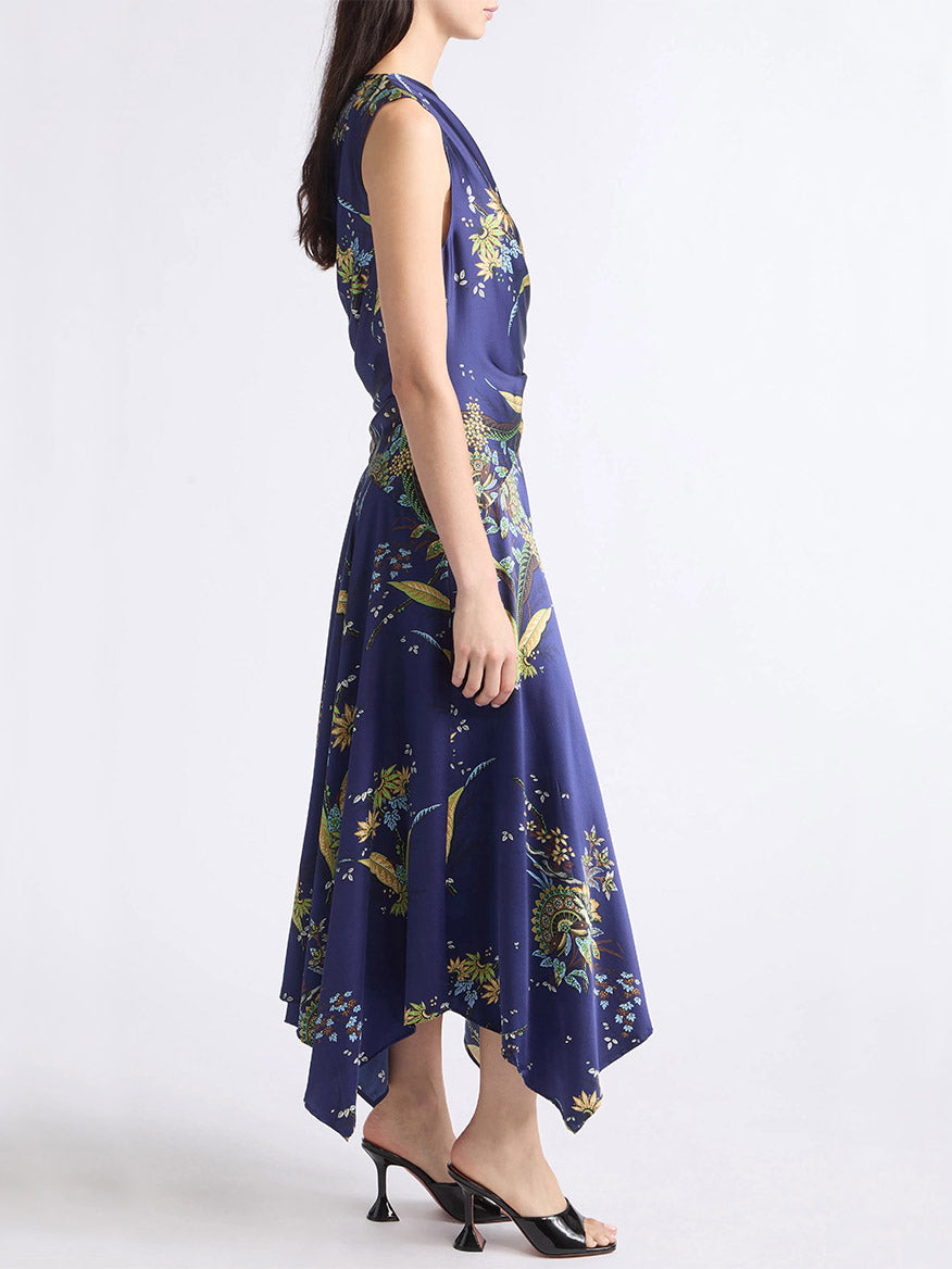 A woman in profile wearing the STAUD Portia Silk Dress in Tapestry Midnight, a flowery printed maxi dress with an asymmetrical hem, paired with black heeled sandals and standing against a plain background.