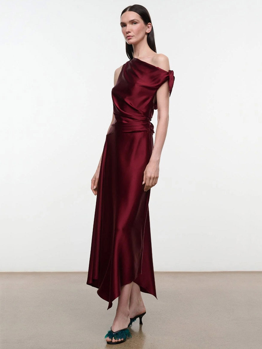 A model wearing the STAUD Portia Silk Dress in Carmino, an asymmetrical burgundy midi dress with a draped bodice and gathered waist with adjustable ties.