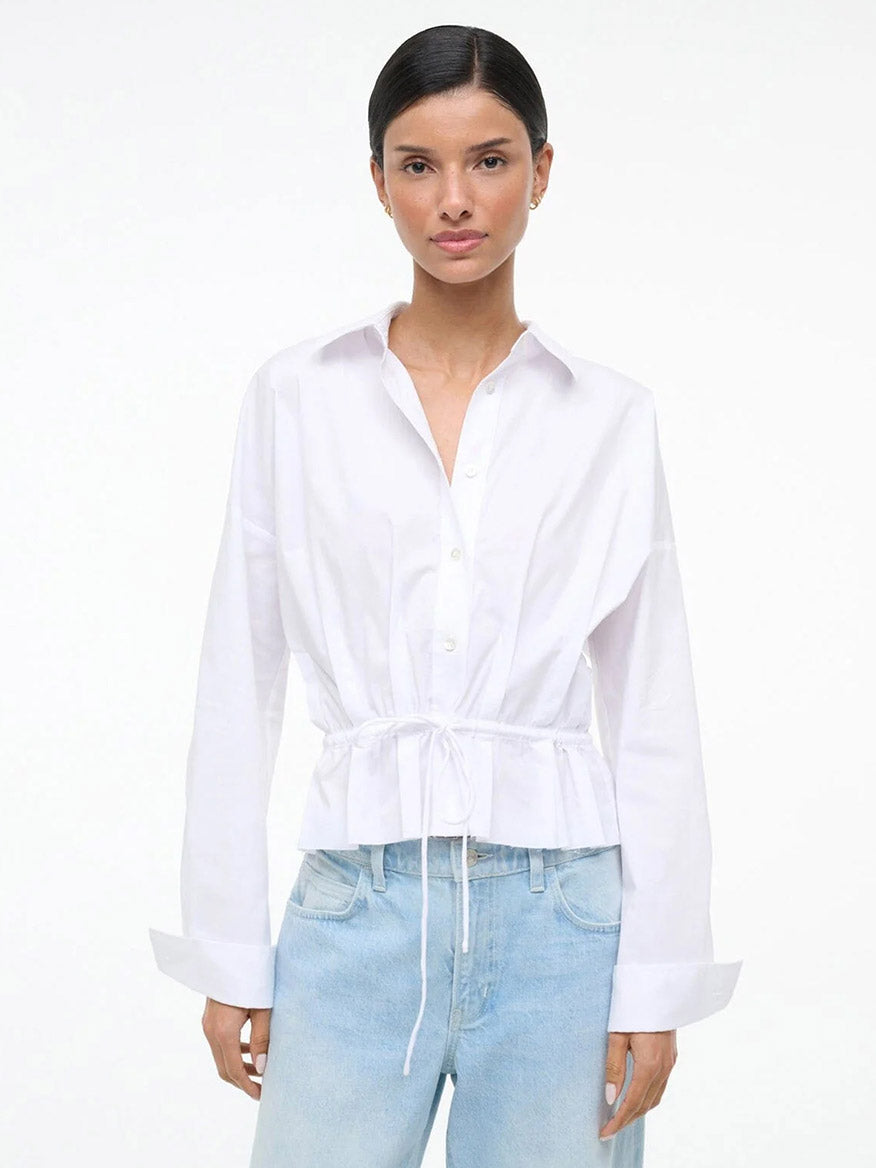 A woman wears the STAUD Primrose Shirt in White styled as a button-down with light blue jeans, standing against a plain white background.