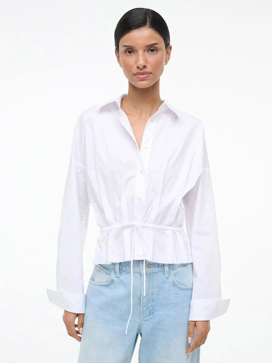 A woman wears the STAUD Primrose Shirt in White styled as a button-down with light blue jeans, standing against a plain white background.