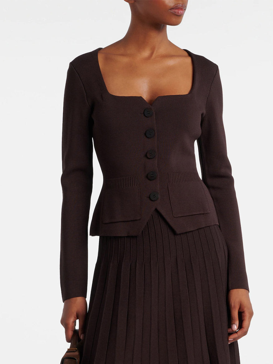 A woman wears the STAUD Rachela Sweater in Earth—a dark brown, long-sleeve knit top with a notched square neck and two front pockets—paired with a matching pleated skirt.