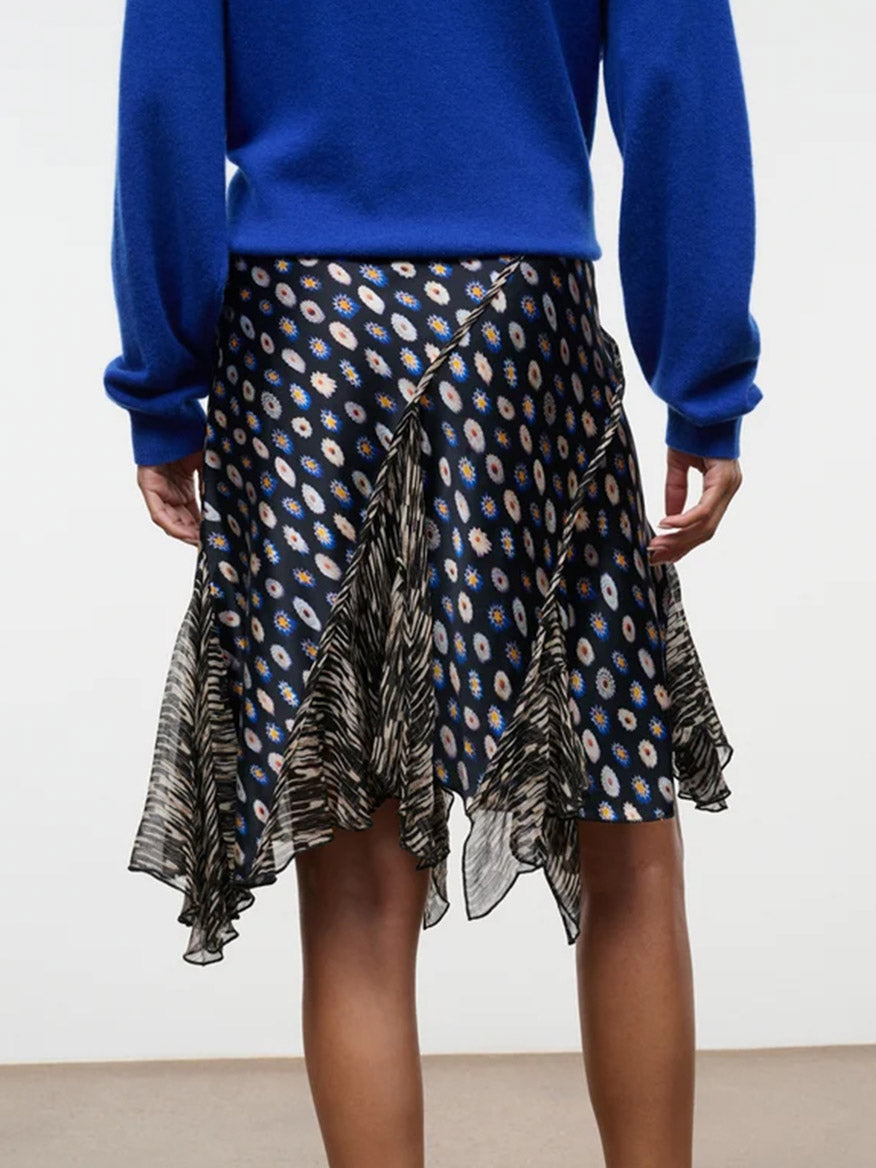 A person wearing a blue long-sleeve top and the STAUD Reannah Silk Skirt in Glasswork Multi, featuring a dark, patterned design with an asymmetric hem and ruffled black and white details, stands facing away.