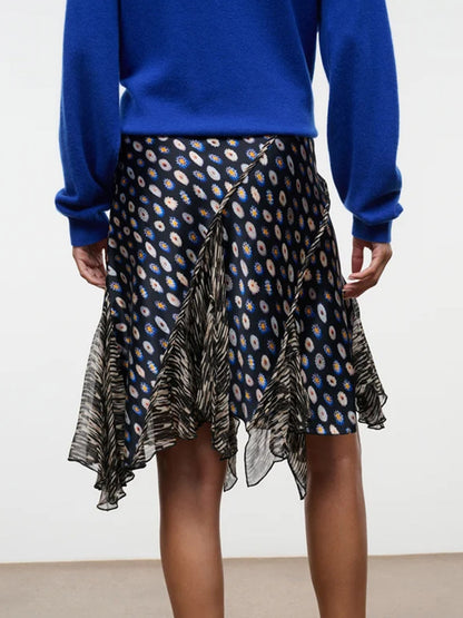 A person wearing a blue long-sleeve top and the STAUD Reannah Silk Skirt in Glasswork Multi, featuring a dark, patterned design with an asymmetric hem and ruffled black and white details, stands facing away.