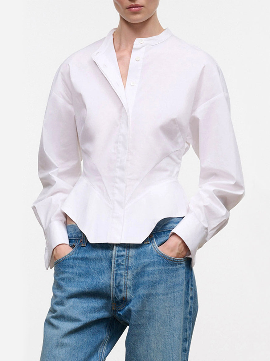 Wearing the STAUD Rhys Stretch Top in White—a band-collar shirt with barrel cuffs, a fitted waist, and a curved hemline—paired with blue jeans, this person stands with hands in pockets against a plain background.