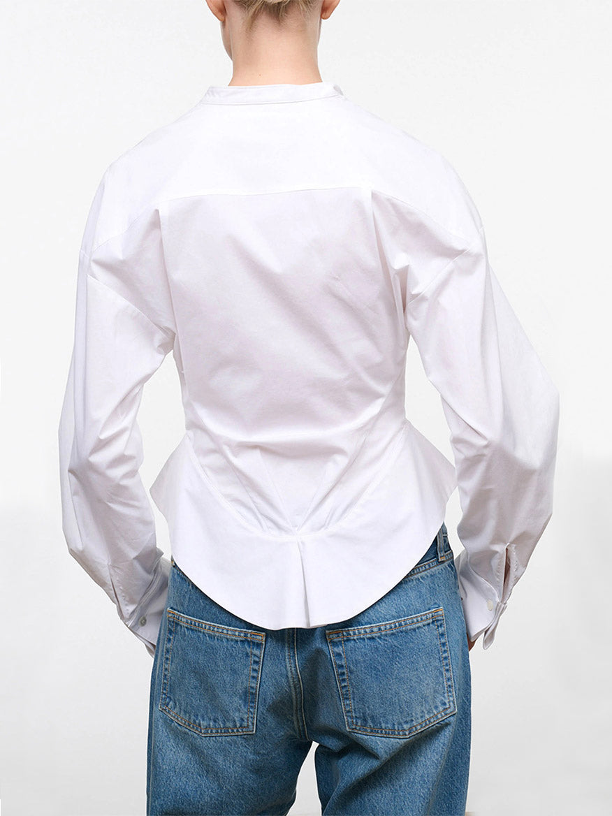 A person is shown from the back against a plain white background, wearing the STAUD Rhys Stretch Top in White—a long-sleeve, band-collar shirt with a curved hemline—paired with blue jeans.