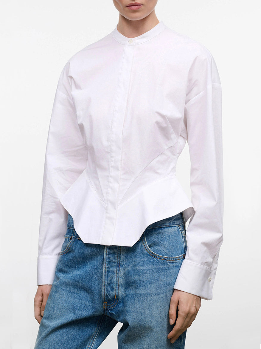 A person wearing the STAUD Rhys Stretch Top in White, a structured long-sleeve band-collar shirt with barrel cuffs and a fitted waist, paired with blue jeans, shown from shoulders to hips against a plain background.