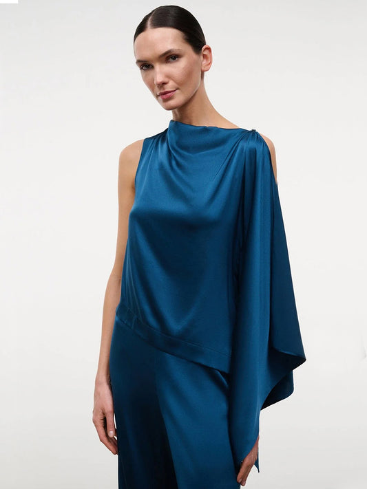 A woman wearing the STAUD Sabina Silk Top in Harbor—a sleeveless, easy-fit stretch silk satin column top with asymmetric draped shoulder detail—stands against a plain white background.