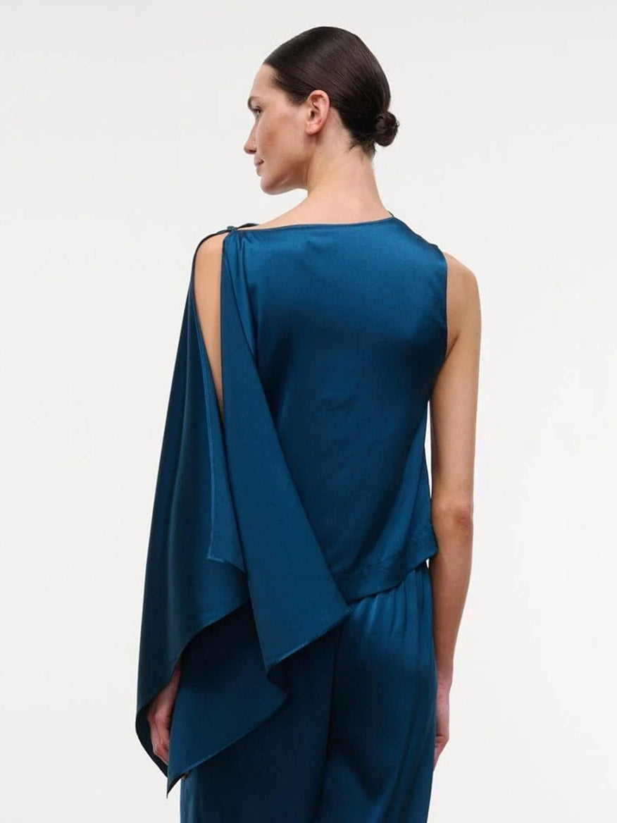 Wearing the STAUD Sabina Silk Top in Harbor, a woman stands facing away. The easy-fit, sleeveless blue stretch silk satin column top features an asymmetrical draped panel over one shoulder and is paired with matching blue pants.