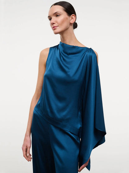 A woman wearing the STAUD Sabina Silk Top in Harbor, an easy-fit, sleeveless asymmetrical stretch silk satin piece with a draped shoulder, styled with matching pants and standing against a plain light background.