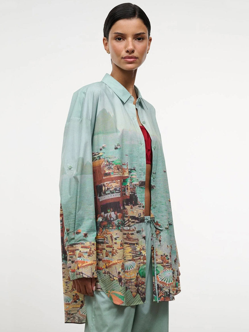 A woman wears the STAUD Sandbar Top in Endless Summer, an oversized button-down with a beach-scene print, layered over a red top and loose green pants against a plain light background.