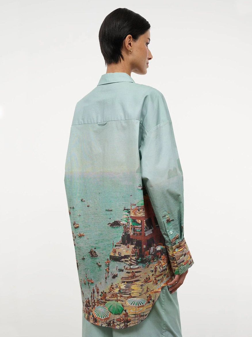 Someone models the STAUD Sandbar Top in Endless Summer, an oversized light green button-down with a detailed beach-scene print on the back, standing against a plain white background.