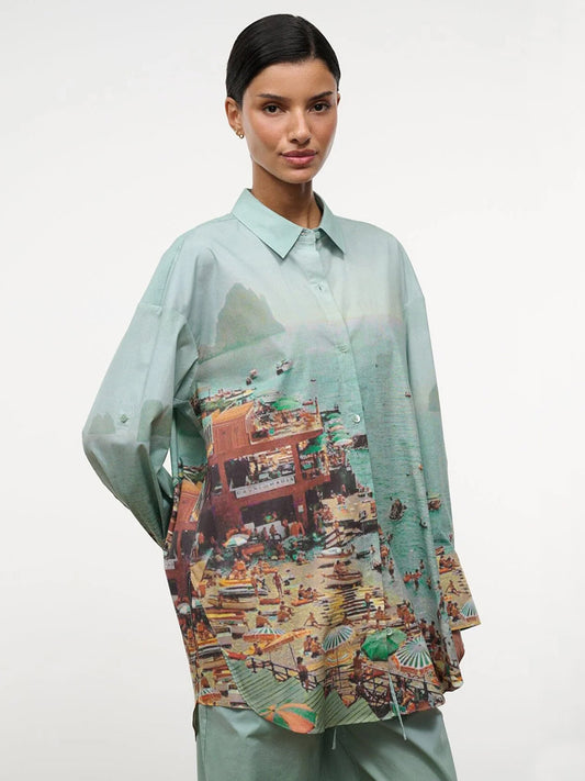 A woman wears the STAUD Sandbar Top in Endless Summer, an oversized pastel green button-down featuring a detailed beach scene with people, umbrellas, and water, standing against a plain white background.