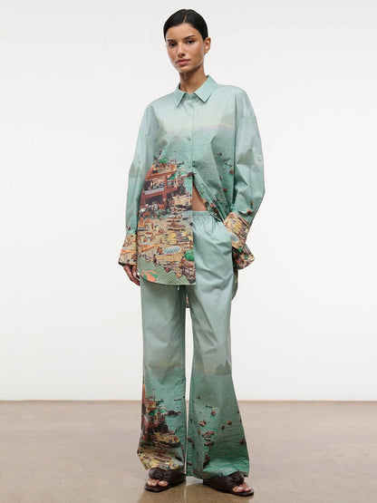 A woman stands against a plain background wearing the STAUD Sandbar Top in Endless Summer, an oversized mint-green button-up shirt with a beach-scene print and matching wide-leg pants.