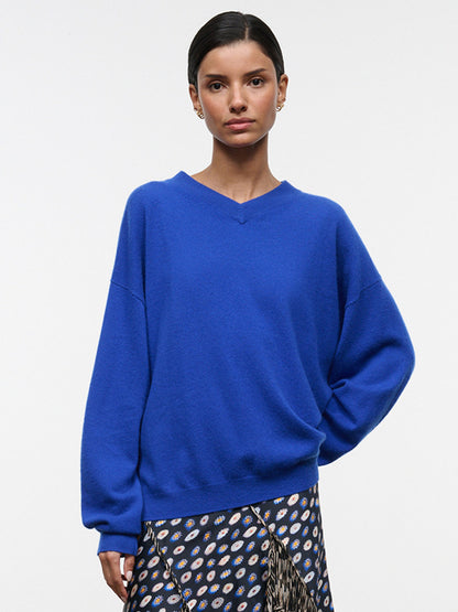 A woman wearing the STAUD Spencer Cashmere Sweater in Cobalt, paired with a patterned skirt, stands against a plain white background, effortlessly showcasing the sweater’s relaxed fit and casual elegance.