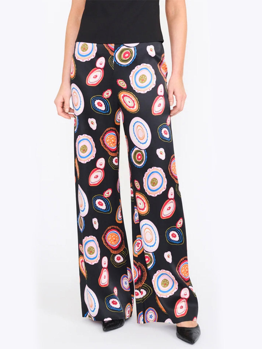 A person wearing the STAUD Stroll Silk Pant in Millefiore 2.0—black wide-leg trousers adorned with a vibrant, circular abstract pattern on a white background, crafted from luxurious stretch silk satin and finished with an elastic waistband for enhanced comfort.
