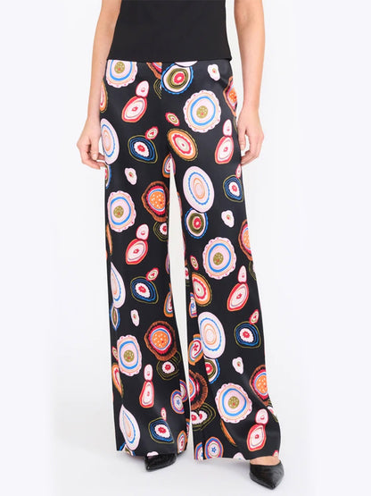 A person wearing the STAUD Stroll Silk Pant in Millefiore 2.0—black wide-leg trousers adorned with a vibrant, circular abstract pattern on a white background, crafted from luxurious stretch silk satin and finished with an elastic waistband for enhanced comfort.
