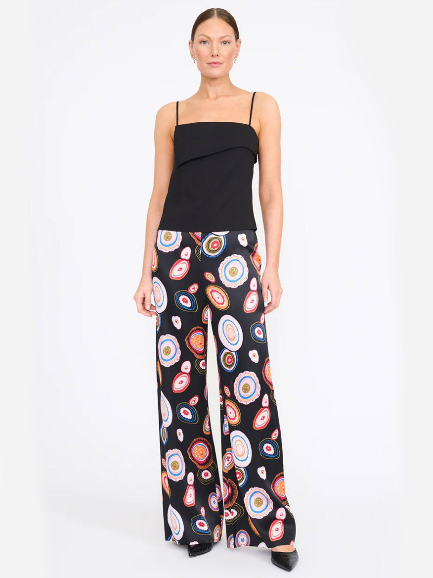 A woman wears the STAUD Stroll Silk Pant in Millefiore 2.0, showcasing its vibrant, colorful circular pattern and elastic waistband, paired with a black spaghetti strap top and standing against a plain white background.
