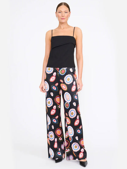 A woman wears the STAUD Stroll Silk Pant in Millefiore 2.0, showcasing its vibrant, colorful circular pattern and elastic waistband, paired with a black spaghetti strap top and standing against a plain white background.