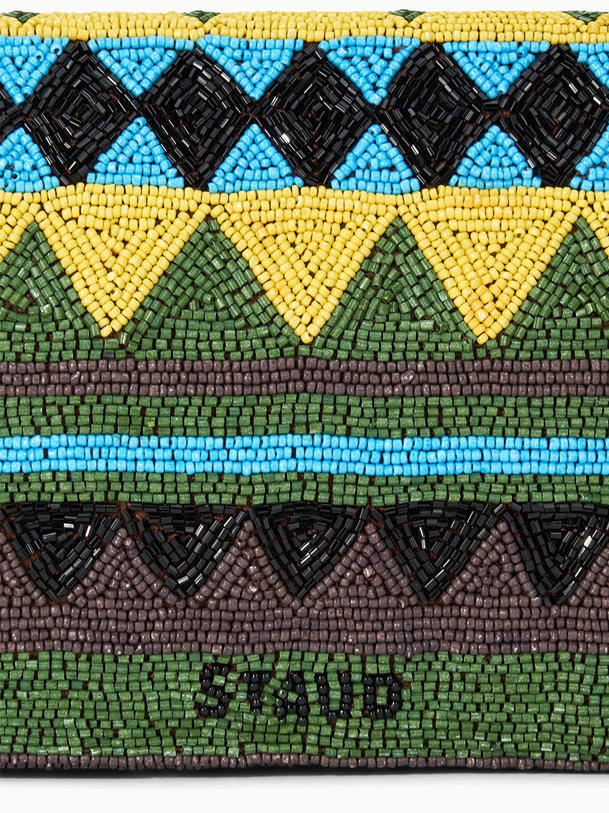 A close-up of the STAUD Timmy Shoulder Bag in Happy Sack, showcasing beaded geometric patterns in black, yellow, green, brown, and blue, with "STAUD" spelled out in black beads at the bottom.